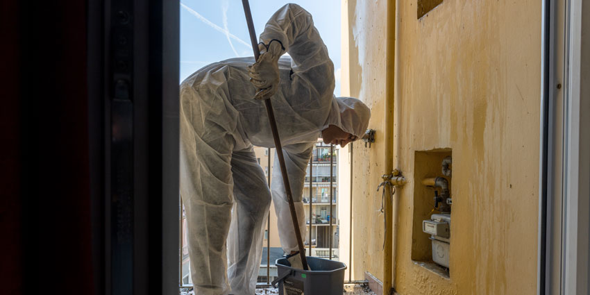 Il professionista della ML Service esegue una disinfestazione a Torino. E' rivestito da una tuta integrale di tessuto non tessuto e indossa guanti protettivi. Lavora su un terrazzino di un condominio e sta ripulendo l'attrezzo utilizzato per la pulizia del muro di colore beige.